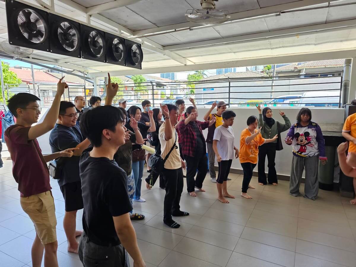 MedicalMet volunteers and residents dancing with arms raised in the open hall of the PKKII Insan home in Happy Garden, Kuala Lumpur